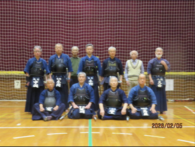 体育館の舞台前で剣道着を着た2人の男性が向かい合い、向かって右の男性は竹刀を振りかざし、向かって左の男性は、右の男性の右わき腹あたりに竹刀をあてようとしている様子の写真