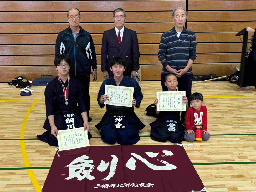 体育館で、手前の床に「剣心」と書かれた赤紫色の応援を広げ、賞状を胸の前に掲げて正面を向き、正座をしている剣道着の男の子2人と、その両脇に座る男の子と女の子、背後に立つ男性3名の集合写真