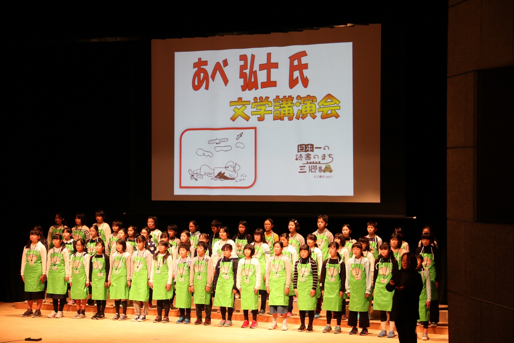 あべ弘士氏文学講演会と書かれた映像が映し出されたスクリーンの下で黄緑のお揃いのエプロンをした子供達が並んでいる舞台を写した写真