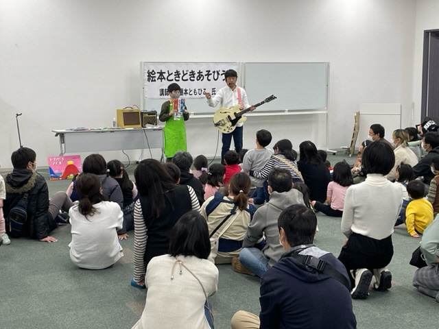 観客の前にギターを持っている男性が立っていてその横に緑色のエプロンをした中学生が本を持っている写真