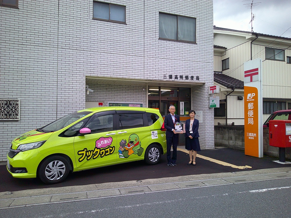 郵便局の前にとめられた黄緑色のラッピングカーの横に人が二人立っている写真