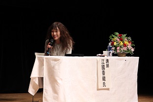 白い布がかけられ花が飾られた机の奥の椅子に座った女性がマイクを持ち話をしている写真