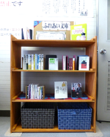 木材の本棚の一番下に籠が2個置かれ、1段目にふれあい文庫の案内、2段目と3段目に本が並べられた彦沢老人福祉センターのふれあい文庫を正面から写した写真