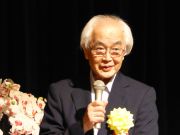 白髪でスーツの胸元に花を飾った男性がマイクを持ち話をしている写真