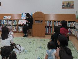 2人の関係者がひよこのイラストが入った紙を見ながら読み聞かせをしており、多くの親子連れの来館者が座って見ている写真