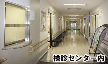 通路右手前にはいくつものパンフレットがパンフレットスタンドに置かれ、右奥にはトイレがあり、左側には黒色の長い待合椅子が置かれている検診センター内廊下の写真